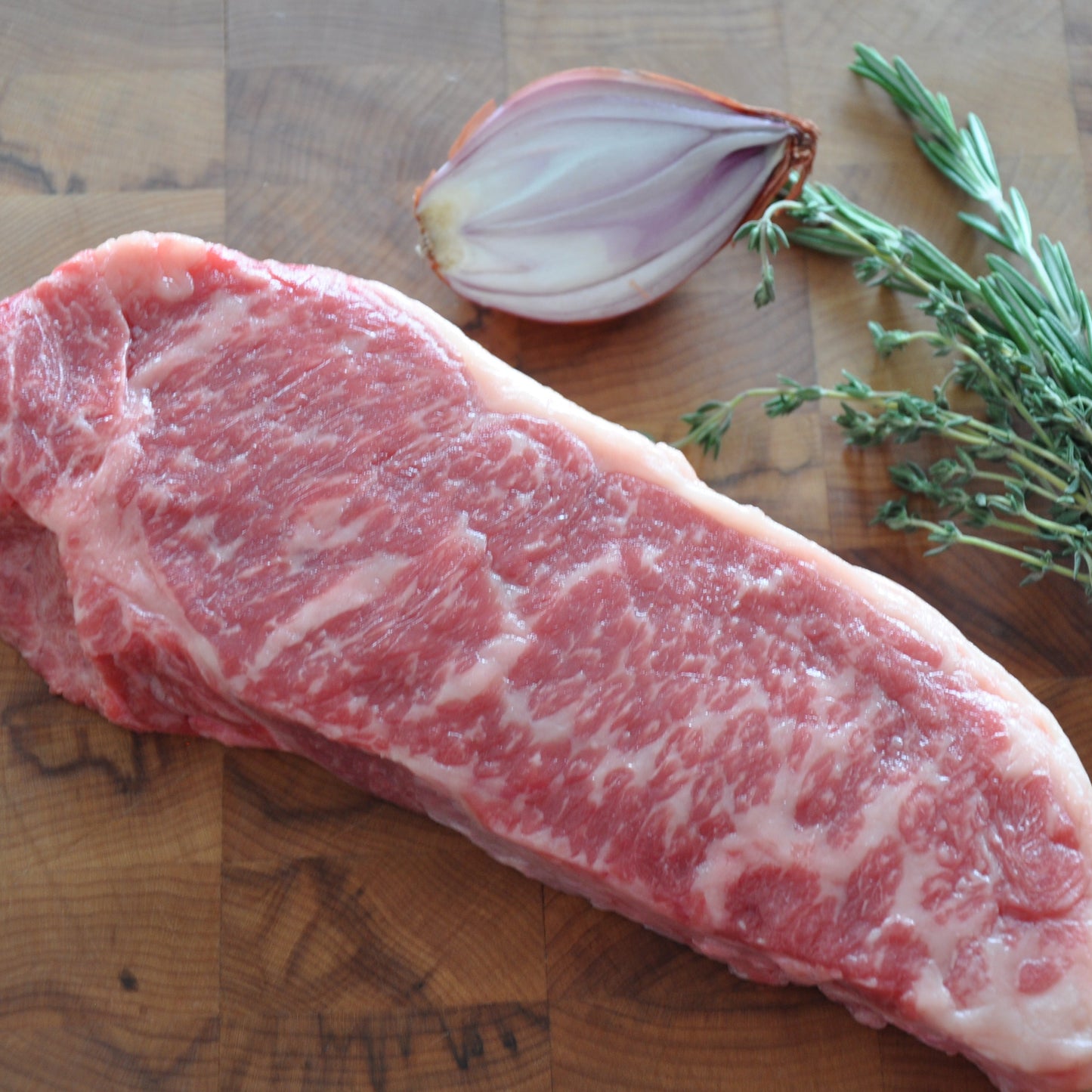 Medium shot of a NY strip cut next to fresh herbs and veggies on top of a wooden board