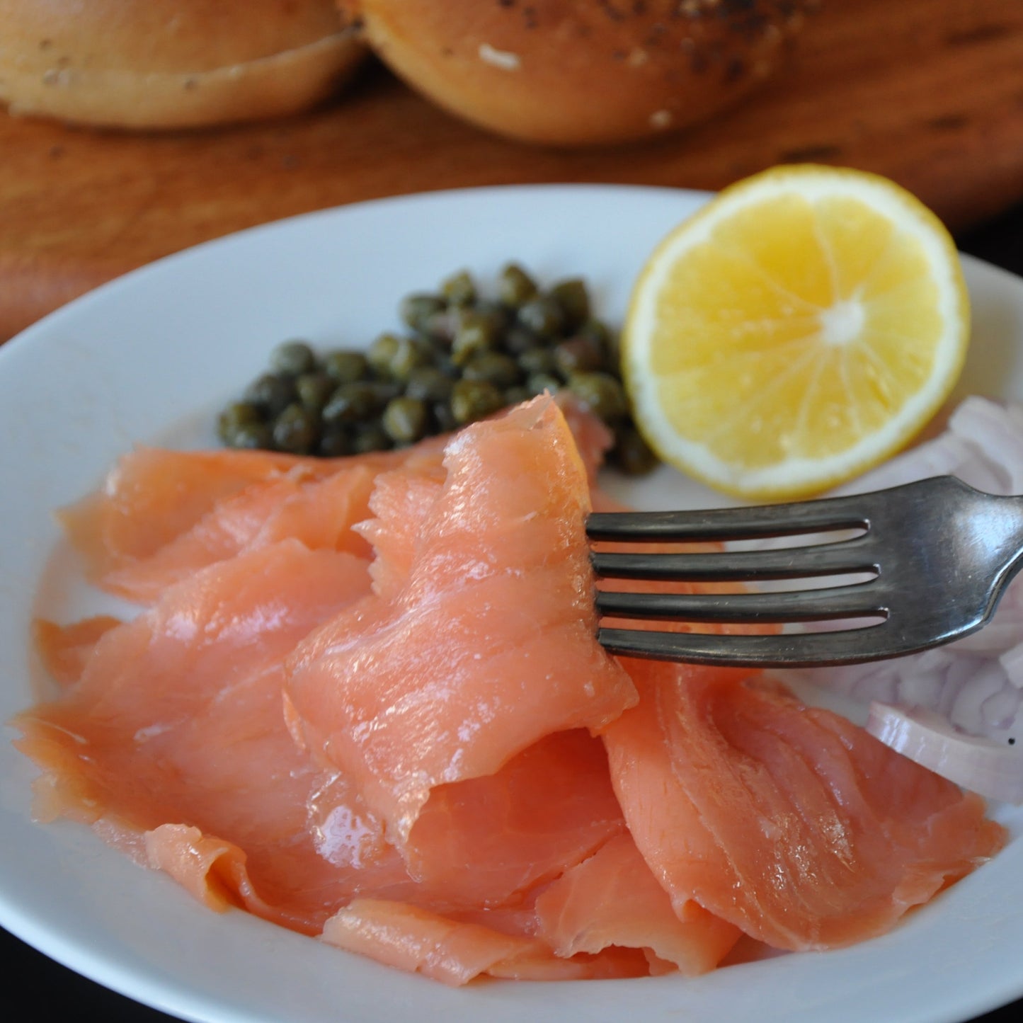 A fork holding a slice of smoked salmon. There's capers and lemon in the background