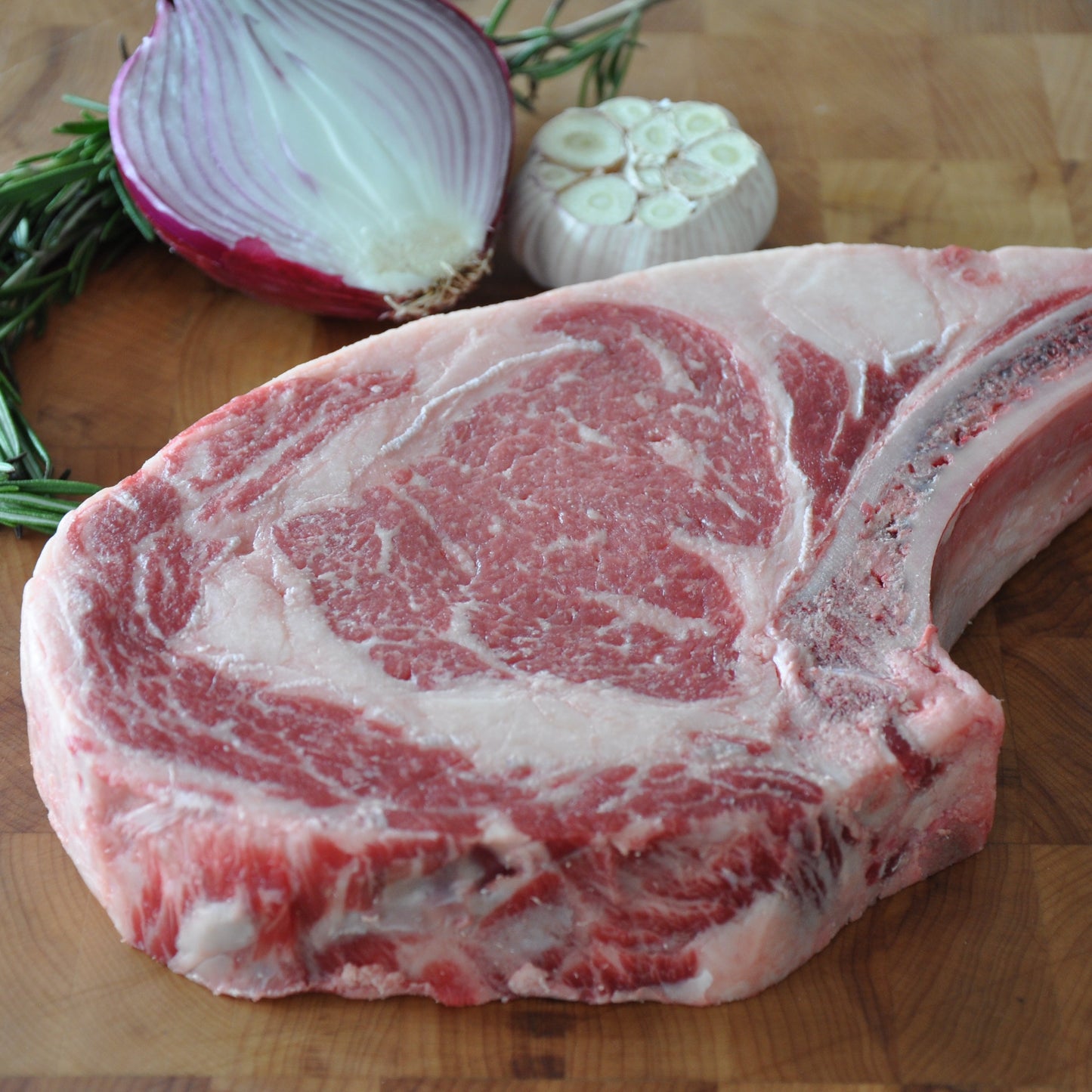 A close up of a bone-in ribeye next to herbs, onion, and garlic on top of a wooden board.