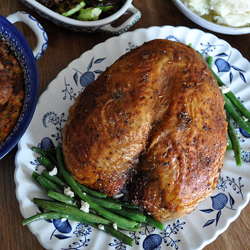 A roasted turkey breast on a white plate with blue floral patterns, served with green beans on a wooden dining table, surrounded by side dishes including stuffing, mashed potatoes, cranberry sauce, and gravy.