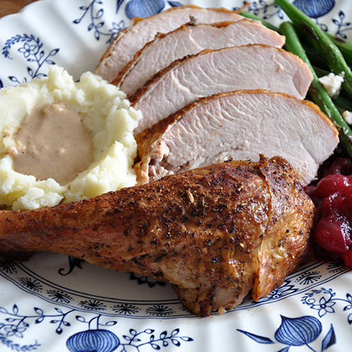 A close-up of a plated Thanksgiving meal featuring sliced turkey, a roasted turkey leg, mashed potatoes with stuffing, green beans, and cranberry sauce on a blue and white patterned plate.