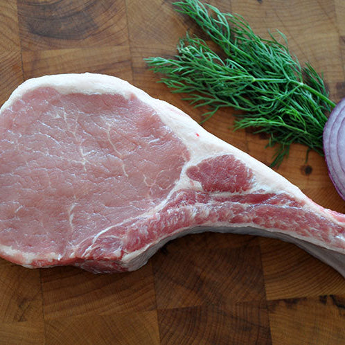 A raw bone-in pork chop next to fresh dill and onion on a wooden board. 