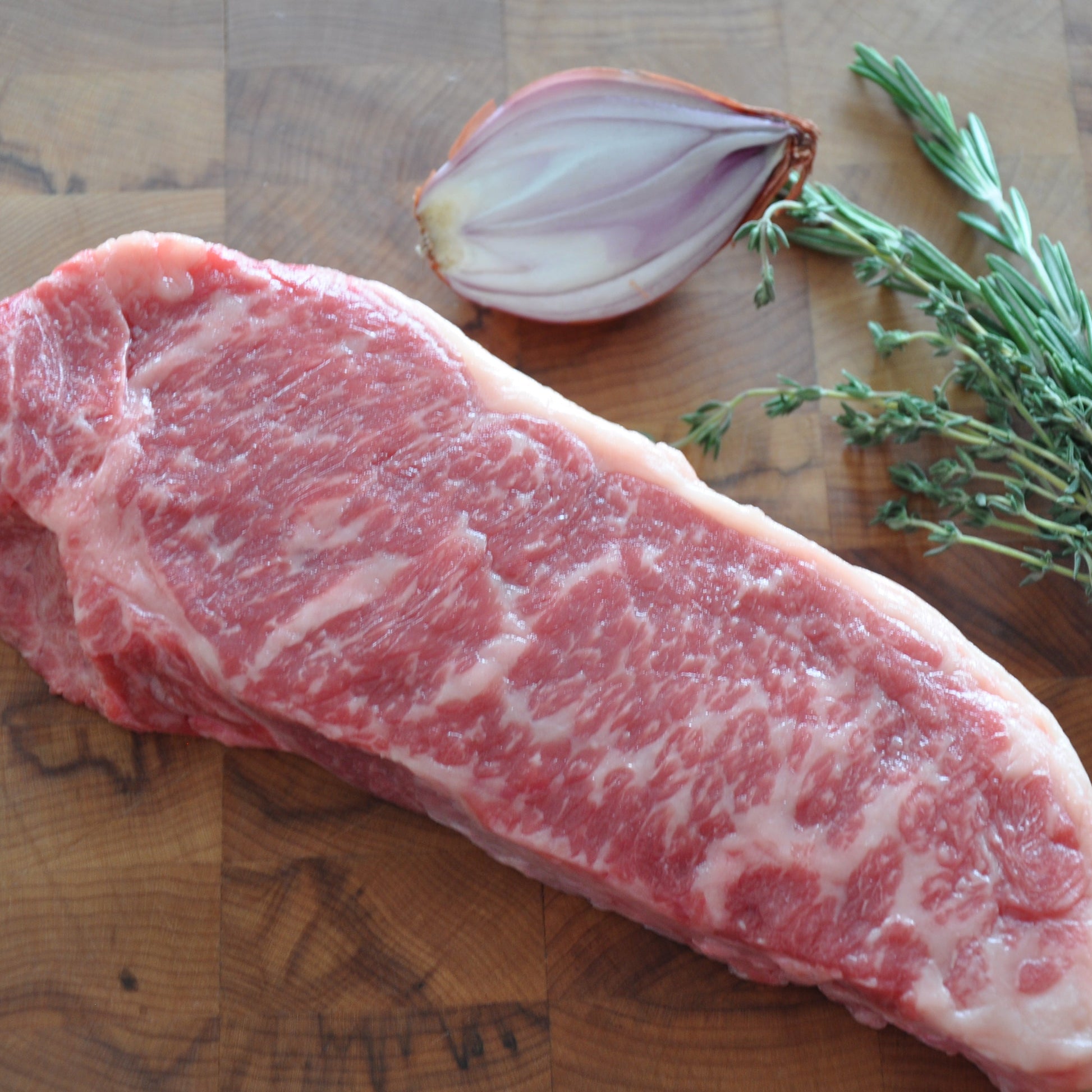 Medium shot of a NY strip cut next to fresh herbs and veggies on top of a wooden board