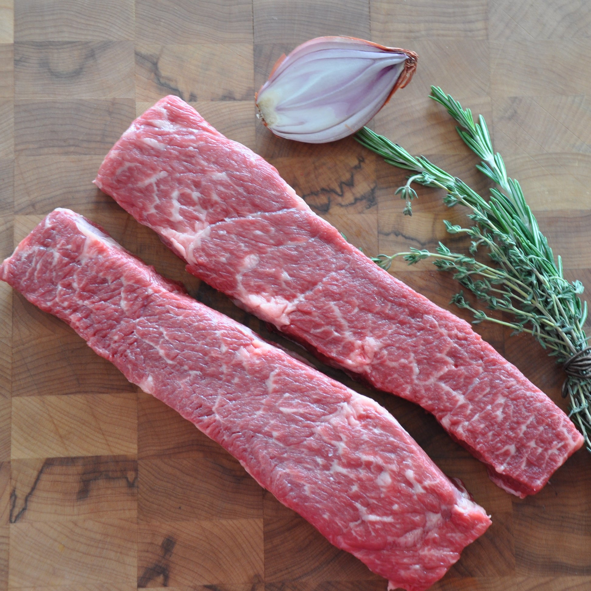 Medium shot of raw prime Denver steaks next to fresh vegetables on a wooden board.