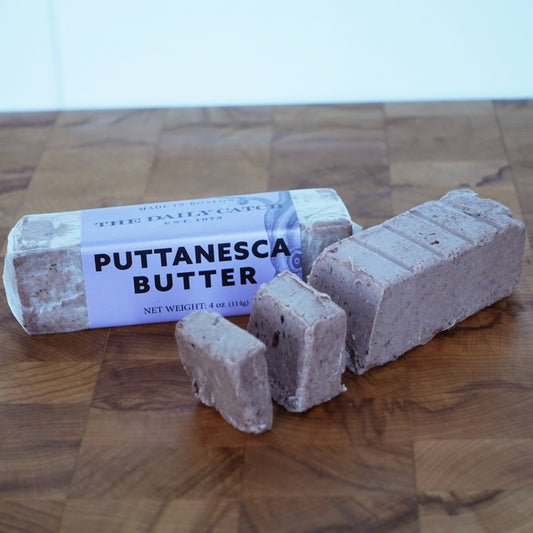 Two butter sticks on a wooden board. The label reads "Puttanesca Butter"