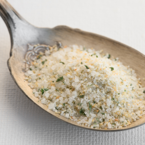 A close-up of a spoonful of coarse seasoning blend containing white salt crystals, green herbs, and orange zest, set against a textured white background.