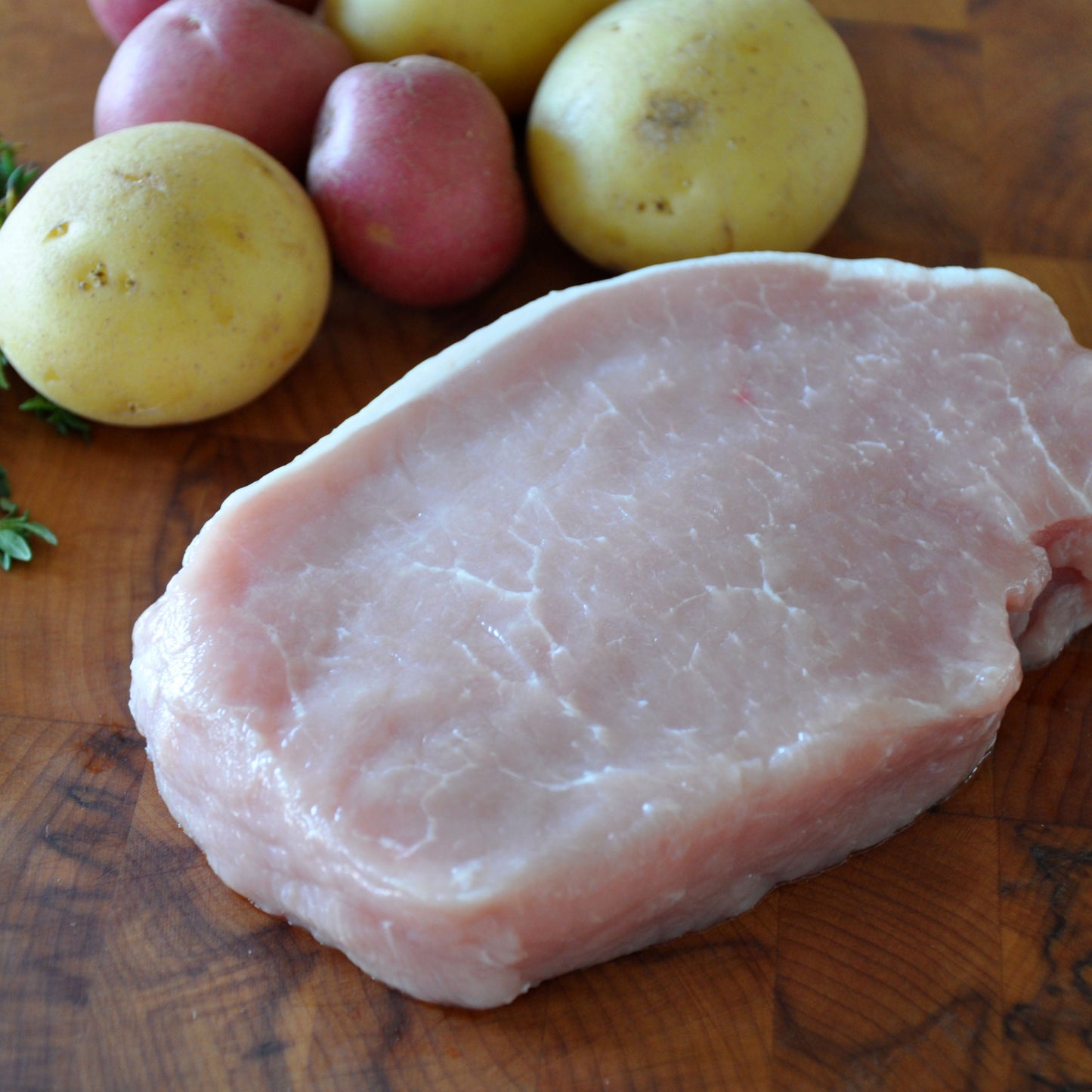 Raw, boneless pork chop next to small potatoes and fresh thyme on a wooden board.