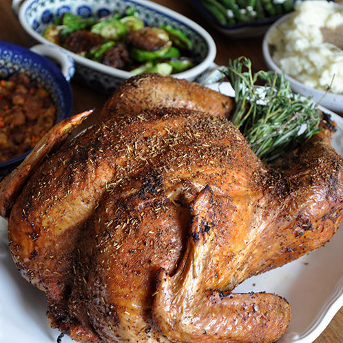 A roasted whole turkey on a white platter, surrounded by holiday side dishes including stuffing, mashed potatoes, green beans, Brussels sprouts, and cranberry sauce on a wooden table.